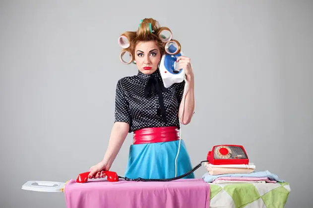 Studio shot of young beautiful housewife by mistake making call with iron and ironing laundry with telephone headphone instead of another way around with gray background behind. Stereotypical housewife have curlers in hair and making confused face