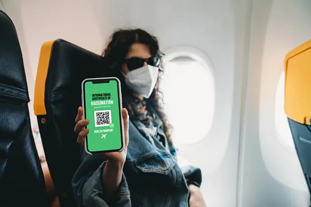 Woman on airplane is wearing a protective face mask and is showing a Covid-19 digital passport to the camera with her smart phone. Green pass with dummy QR-code on the phone screen.