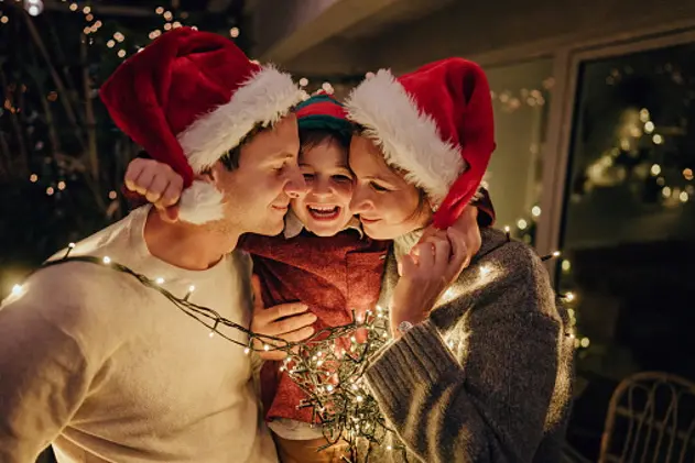 Photo of a family with one child wrapped in Christmas lights,  celebrating holidays on the balcony of their apartment; throwing an unforgettable Christmas party at home.