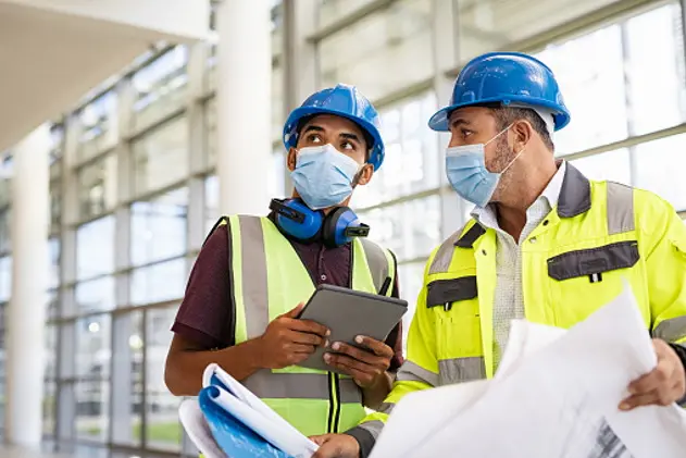 Mixed race architect and technician at construction site reviewing blueprints while wearing surgical face mask and hardhat. Team of specialists construction worker with face mask using digital tablet with plans. Young indian architect with mature engineer wearing hard helmet and protective mask for safety against coronavirus while working.