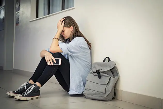 Upset and depressed girl holding smartphone sitting on college campus floor holding head. University sad student suffering from depression sitting on floor at high school. Lonely bullied teen in difficulty with copy space.