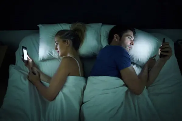 Shot of a young couple using their cellphones in bed at night back to back