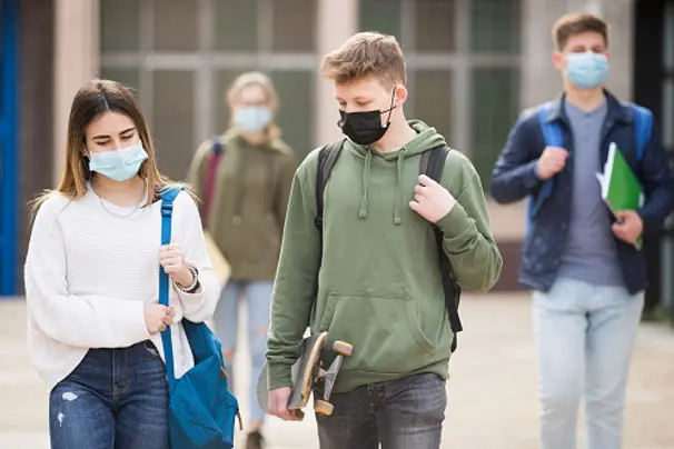 Teen girl in protective face mask walking with her classmate outside college building on sunny day