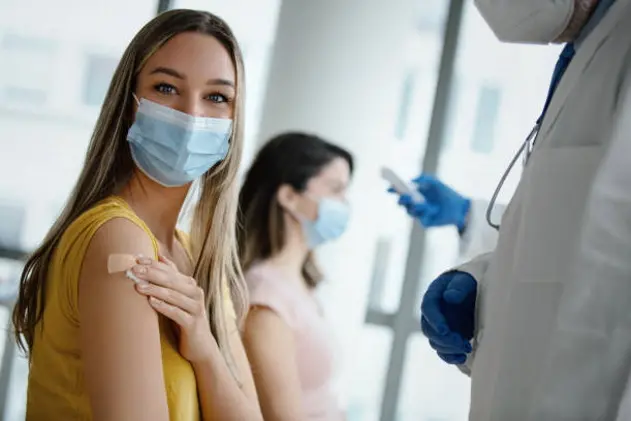 Doctor had just vaccinated a young female patient in the hospital. She is pleased with how it all went. She\\'s smiling behind a protective face mask and looking at camera. Medical worker is in protective workwear.