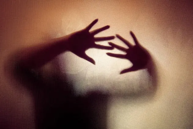 Colour backlit image of the silhouette of a woman with her hands pressed against a glass window. The silhouette is distorted, and the arms elongated, giving an alien-like quality. The image is sinister and foreboding, with an element of horror. It is as if the \\'woman\\' is trying to escape from behind the glass. Horizontal image with copy space.