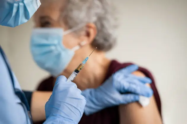 Close up of general practitioner hand holding vaccine injection while wearing face protective mask during covid-19 pandemic. Young woman nurse with surgical mask giving injection to senior woman at hospital. Close up of nurse holding syringe to vaccine old patient from covid19.