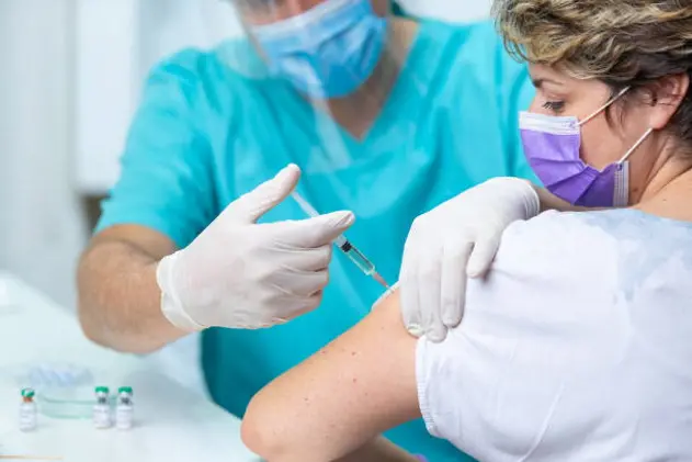 Female patient with protective face mask looking at her arm while doctor in protective gloves is injecting flu vaccine