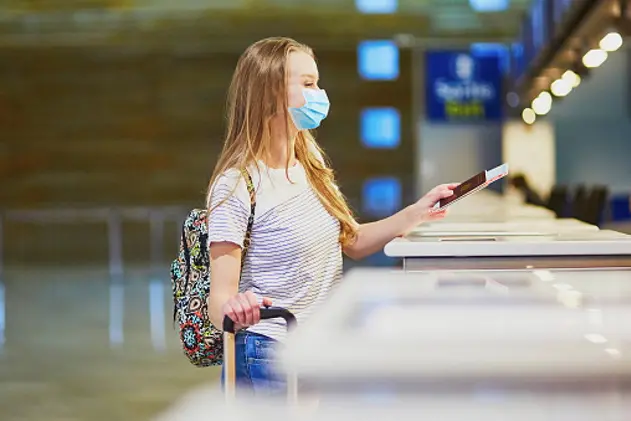 Girl wearing protective face mask with backpack in international airport at check-in counter, giving her passport to an officer and waiting for her boarding pass. Traveling during pandemic