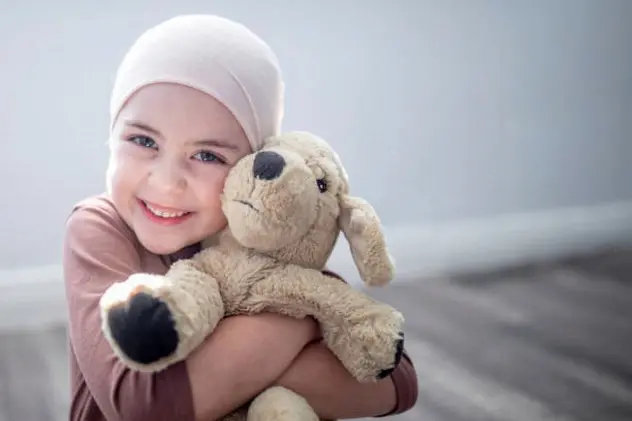 A young girl who has leukaemia is sitting on the floor in her room. She is wearing a headscarf due to her hair loss and is holding onto her favourite teddy bear. She is smiling widely at the camera.