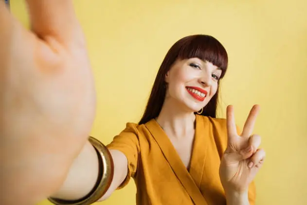 Smiling brown-haired pretty girl in good mood, with bright make up, red lipstick, wearing yellow shirt and bracelets, posing making selfie on bright yellow background