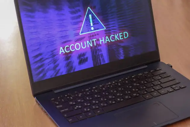 Laptop on the table and on a blue screen the text account is hacked. Warning triangular sign with exclamation mark symbol. Horizontal.