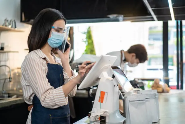 Asian cafe business owner wear protective face shield and mask due to Covid 19, use phone talk to customer and check order from touch screen on counter bar. Waiter prepare food for pick up, takeaway.