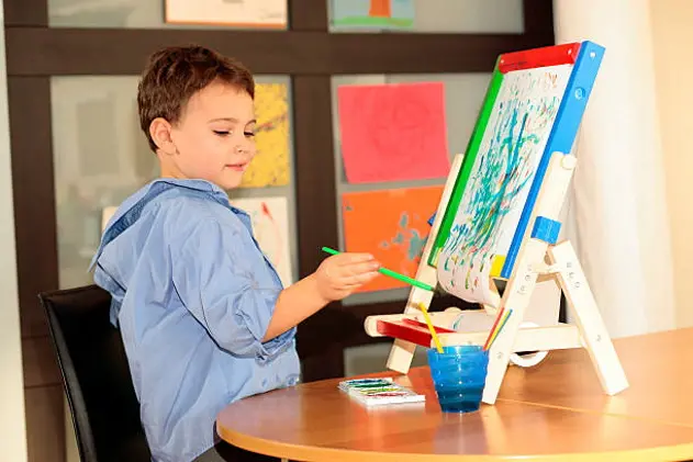 Young boy putting the finishing touches on his watercolor painting. Hanging artwork on the wall behind him are all his creations.