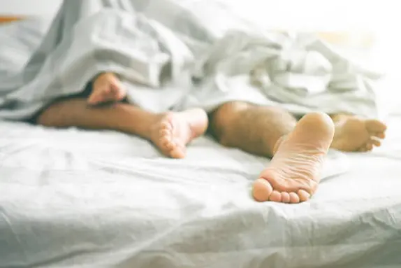 Close up of male and female feet on a bed - Loving couple having sex under under white blanket in the bedroom - Concept of sensual and intimate moment of lovers - Vintage filter - Focus on male foot