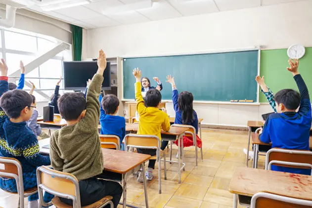 A rear view photo of a group of male and female elementary school children while raising their hands to ask or answer questions in an elementary school building classroom.