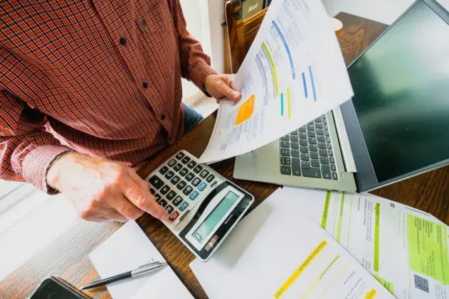 Wide angle image depicting a senior man\\'s hand holding an energy bill while the other hand checks the numbers on a calculator. The table is strewn with documents and also a laptop. Room for copy space.