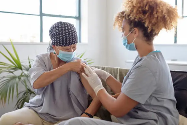 A senior woman of Asian descent with cancer sits on a sofa and lifts the sleeve of her shirt up so a nurse can give her the COVID-19 vaccine. Both women are wearing protective face masks.