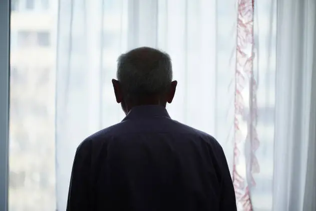 Silhouette of retired man looking through window with transparent curtain standing at home rear view. Loneliness and old human care concept.