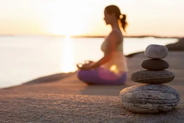 Side view of woman sitting in lotus position on lakeshore with focus on stack of stones