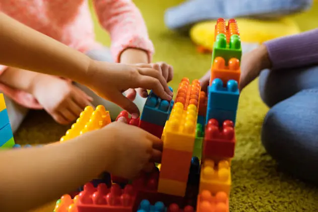 Close up of two unrecognizable kids building something with plastic blocks on carpet at playroom.