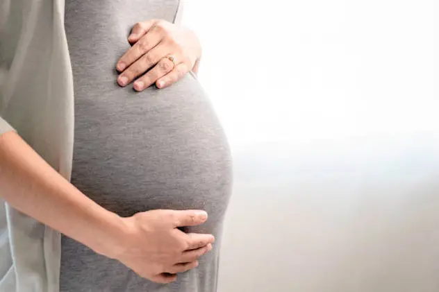 Image of A woman pregnant in gray maternity dress standing near the window. Mom is touching and caressing her belly with her hands. Motherhood, pregnancy, gynecology and health care concept.