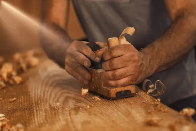 The wood artisan, a robust and strong man known as a carpenter, hand planes a board in the traditional manner, producing shavings of sawdust that look like golden curls of wood. It is a strenuous and