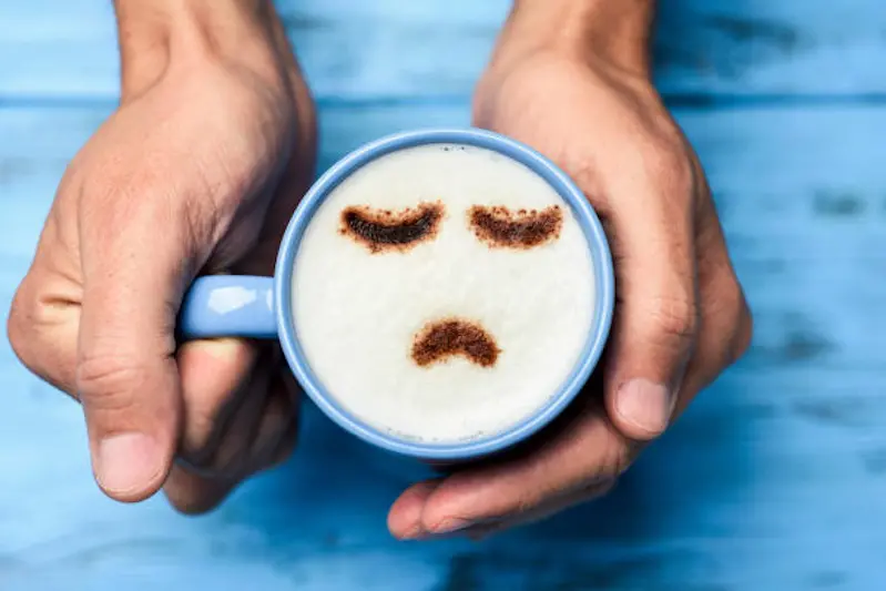 high-angle shot of a young caucasian man with a blue cup of cappuccino with a sad face drawn with cocoa powder on the milk foam, on a blue rustic table