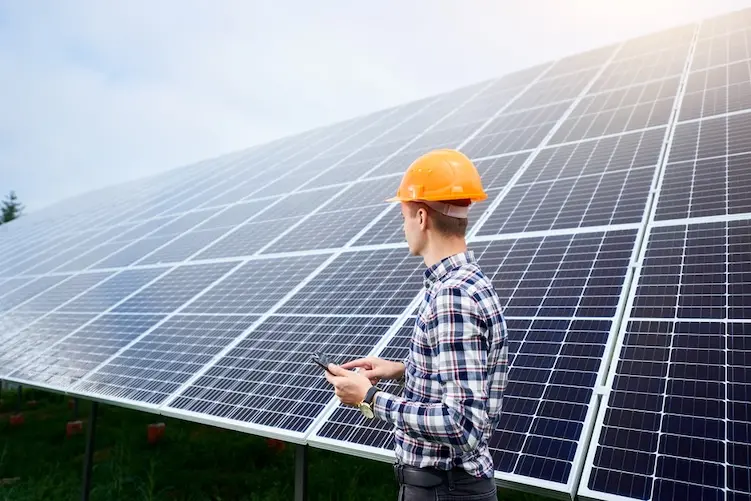 Engineer in helmet with tablet in hands standing near solar panels on green plantation. Concept ecology protection. Man worker. Plant industry.