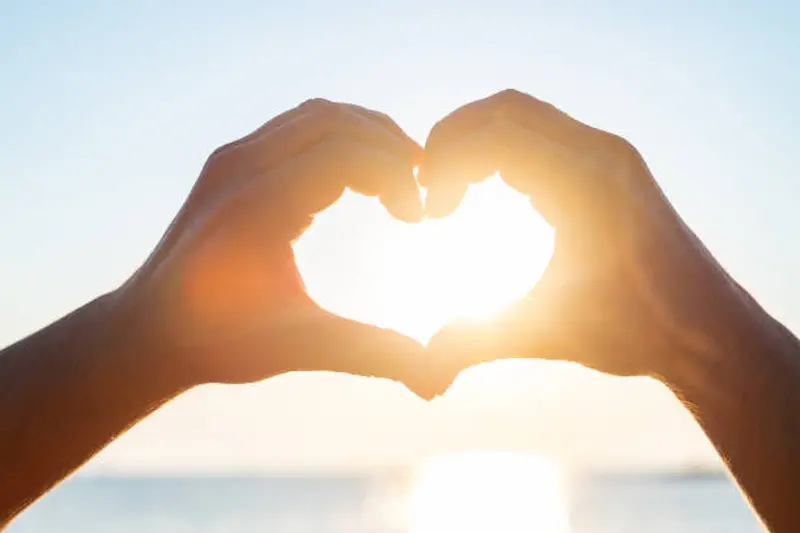 Human hands making shape of heart in front of sun