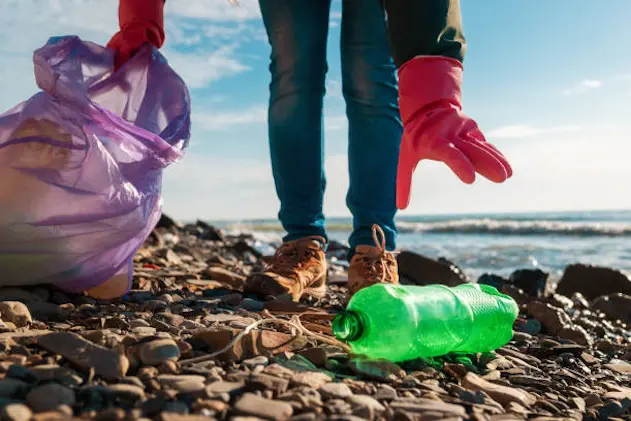 A volunteer in rubber gloves reaches for a dirty plastic bottle lying on the ocean shore. Hand close-up. The concept of clean up of wild beach.