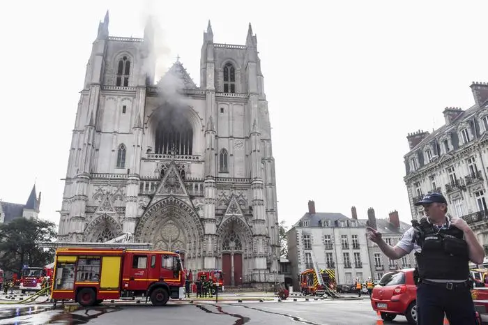 A French Police officer gestures as firefighters are at work to put out a fire at the Saint-Pierre-et-Saint-Paul cathedral in Nantes, western France, on July 18, 2020. - The major fire that broke out on July 18, 2020 inside the cathedral in the western French city of Nantes has now been contained, emergency services said. \"It is a major fire,\" the emergency operations centre said, adding that crews were alerted just before 08:00 am (0600 GMT) and that 60 firefighters had been dispatched. (Photo by Sebastien SALOM-GOMIS / AFP)