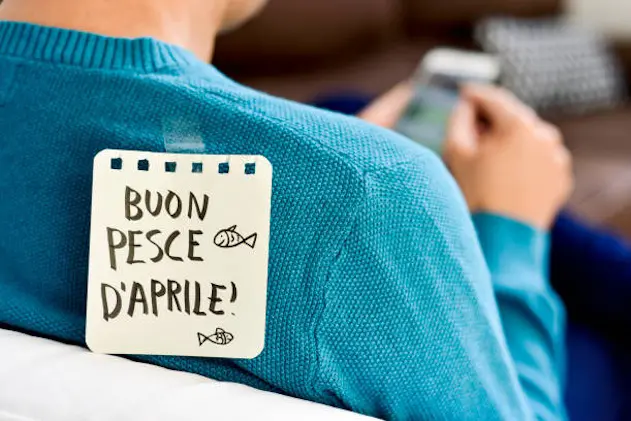 closeup of a young caucasian man seen from behind using his smartphone, with a note with the text buon pesce d aprile, happy april fools day in italian, attached with tape to his back