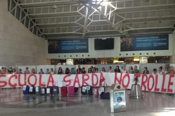 \"Se andiamo a insegnare fuori dalla Sardegna chistionausu in limba, ovvero parliamo in sardo\". \\u00BB il messaggio dei docenti, che da giorni combattono per non emigrare, durante un flash mob all\\'aeroporto di Cagliari-Elmas. Qualche insegnante donna ha indossato un costume tipico di Quartu, altri hanno portato un bronzetto nuragico. E\\' stao anche mostrato un maxi striscione con la scritta \"Scuola sarda no trolley\". Domani alle 14 scade il termine per presentare le domande di assunzione. Cagliari, 13 agosto 2015. ANSA/STEFANO AMBU\\n\\n\\n