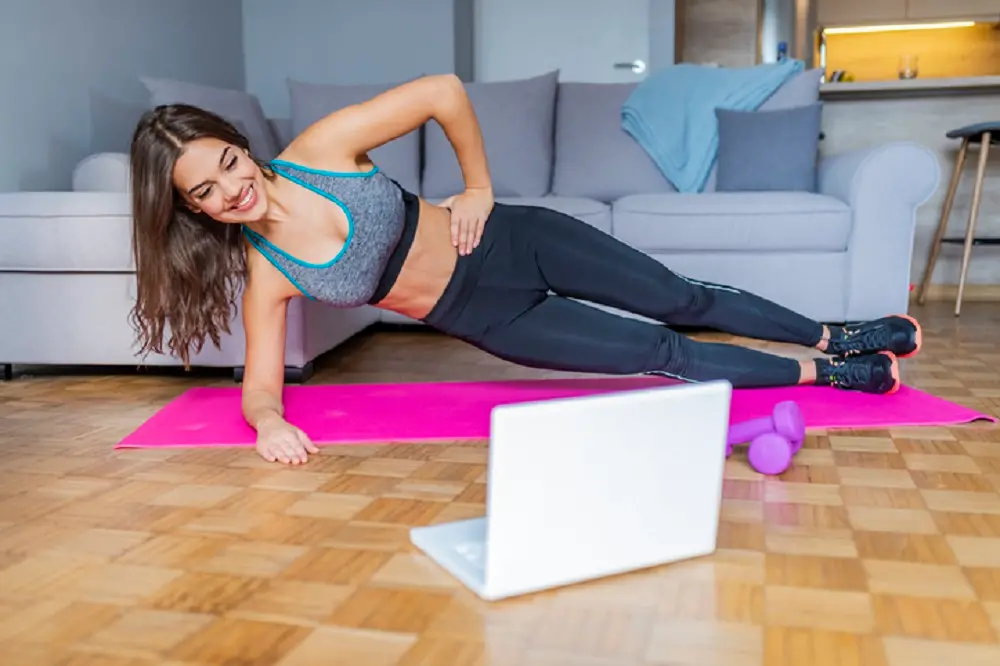 <p>Young woman exercising at home in a living room. Video lesson. Young woman repeating exercises while watching online workout session. Beautiful young woman doing fitness exercise at home</p>\\n